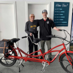 Two happy riders stand next to a Pedego tandem ebike. Indoor photo.